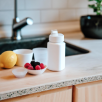 A kitchen countertop with healthy ingredients: berries, yogurt, green tea, lemon water, and a clean white supplement bottle. Bright, crisp lighting.