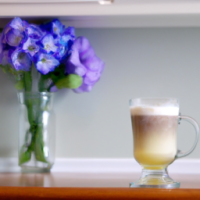 A picture of  a steaming vanilla latte in a clear mug,  setting on a kitchen table surrounded by a small vase with blue flowers