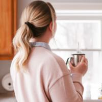 a woman holding a cup of vanilla latte while standing in her kitchen gazing out the window
