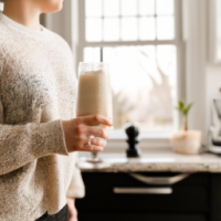 a close up of a woman holding a tall glasss of vanilla latte while standing in her kitchen gazing out the window