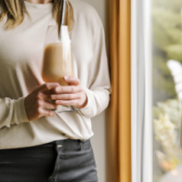 a close up of a woman holding a tall glasss of vanilla latte while standing in her kitchen gazing out the window