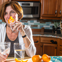 A woman in her early 50's drinking a glass of ice water with orange peels in them. On the kitchen counter has a bowl of oranges on it.