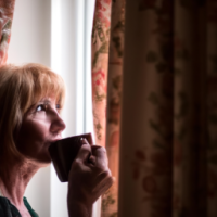 a english women in 50's drinking coffee -looking curios Standing in a window with a soft golden light . curtain open to reveal the morning light . want the theme to be the most powerful part of day 