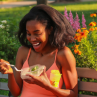 A BEAUTIFUL BLACK WOMAN EATING A SALAD AND DRINKING WATER ON A PARK BENCH SMILING WHILE LOOKING AT BEAUITFUL FLOWERS YOU