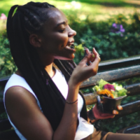 A BEAUTIFUL DARK SKINNED BLACK WOMAN WITH BRAIDS IN HER HAIR WITH A HEALTHY APPEARANCE EATING A SALAD AND DRINKING WATER ON A PARK BENCH SMILING WHILE LOOKING AT BEAUITFUL FLOWERS YOU