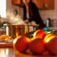 A pot overflowing with boiling orange peels in a kitchen with vegetables on the bench and a woman preparing food in the background