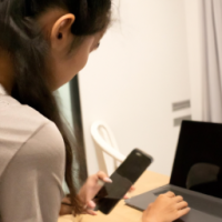 An over the shoulder view of an attractive 26 year old Thai woman typing on her laptop with a mobile phone on the side